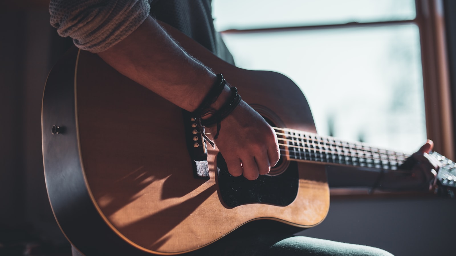 A guitarist playing a steel-string acoustic guitar in warm window light